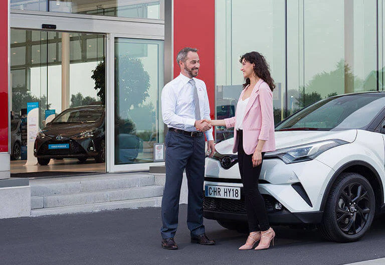 En kvinna och en man som tar i hand framför en Toyota och en Toyota-anläggning.